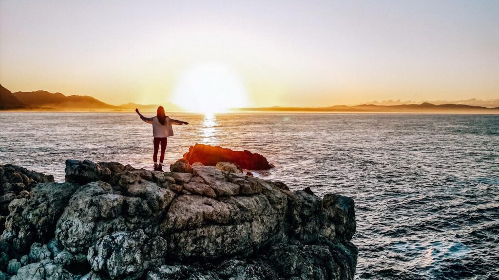 Woman standing on rocks next to the ocean at sunset on a walking trail in Hermanus